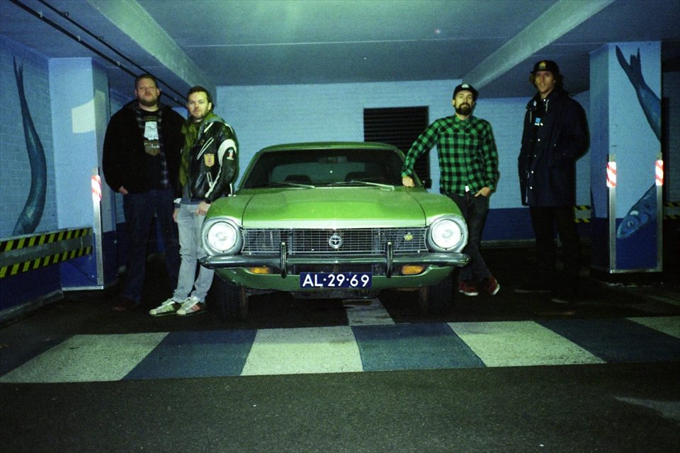 Snowburner band photo: four band members around a green vintage car in a parking garage